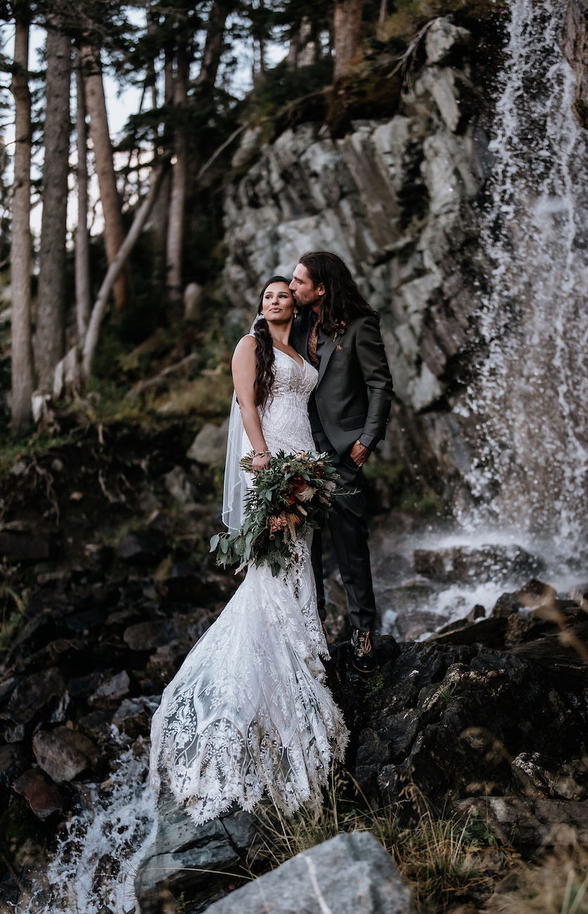 A bride and groom stand together in wedding attire beside a rocky waterfall in a forested area, with the groom kissing the bride on the forehead.