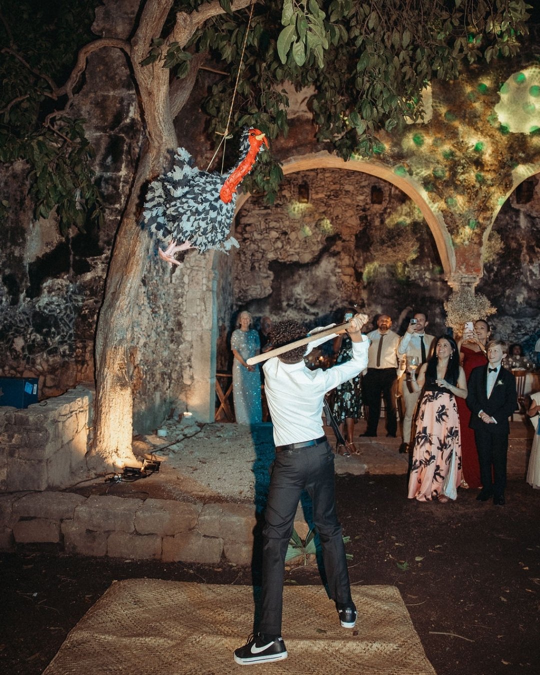 A person swings a bat at a suspended piñata outdoors at night while a group of people in formal attire watch.