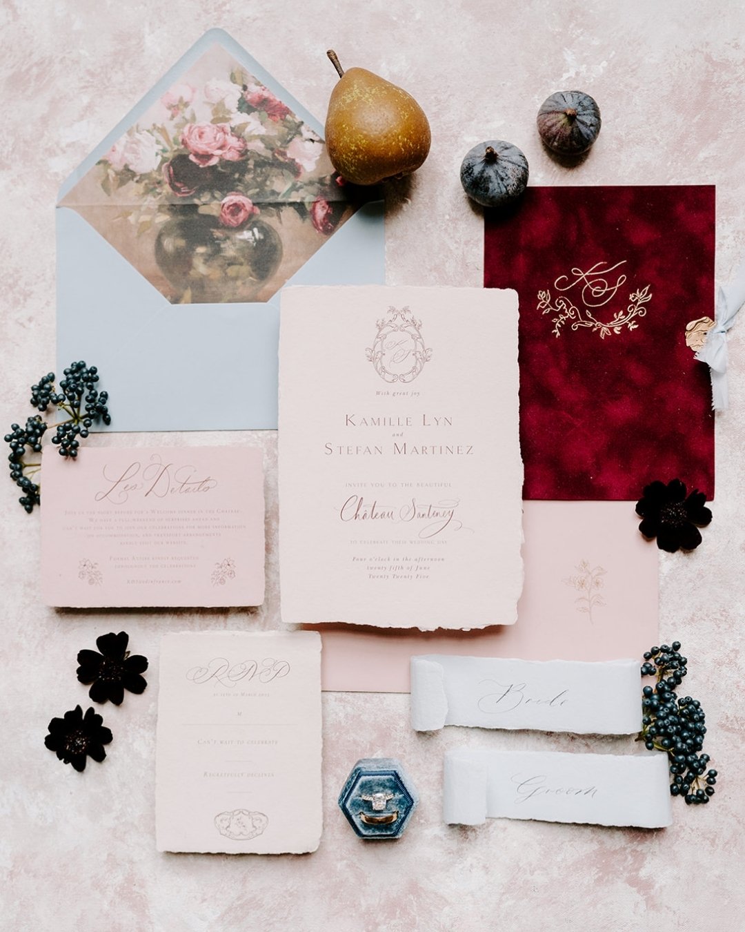 A flat lay of elegant wedding stationery, including invitations, envelopes, a pear, figs, dark flowers, and a red velvet piece, all arranged on a light textured surface.