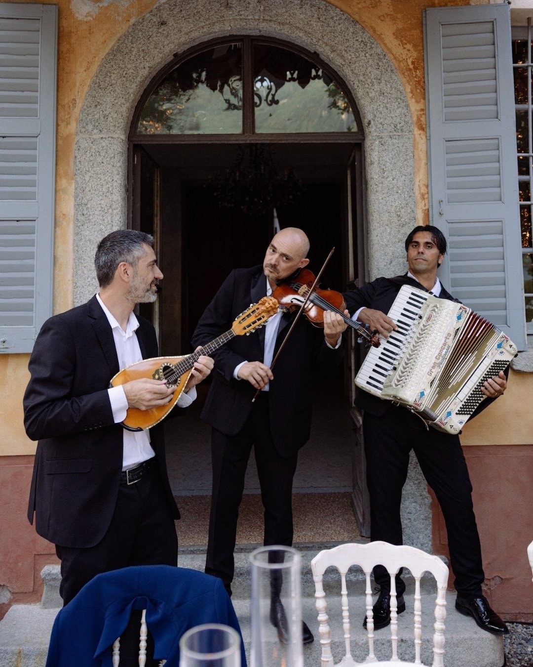 Three men in suits play a mandolin, violin, and accordion outside a building with open shutters and a doorway behind them.