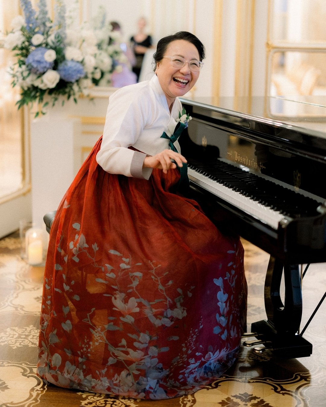 A woman wearing a traditional Korean hanbok sits at a grand piano, smiling broadly in an elegant room with floral arrangements.