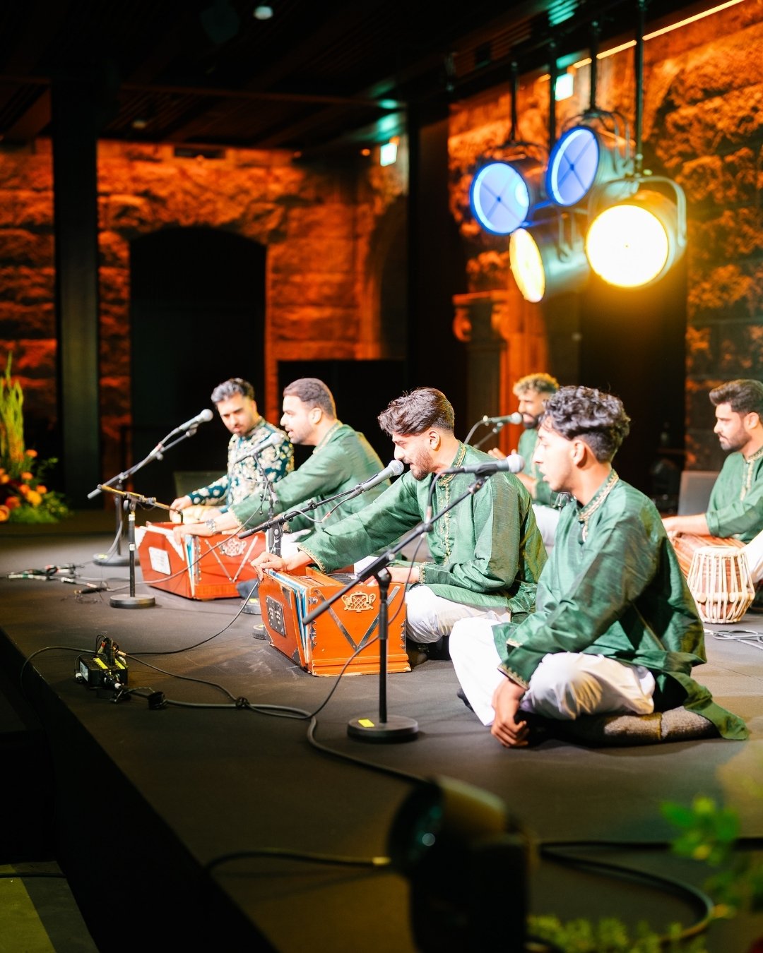 A group of musicians in green outfits sit cross-legged on stage, playing harmoniums and tabla, with microphones and stage lights above them.