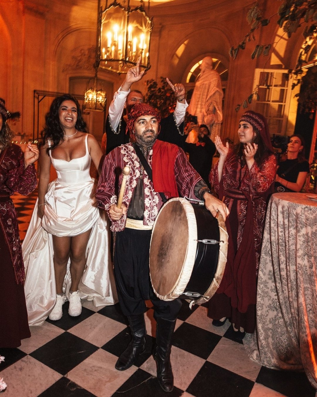 A man in traditional clothing plays a large drum at an indoor event as others, including a woman in a white dress and sneakers, clap and celebrate around him.