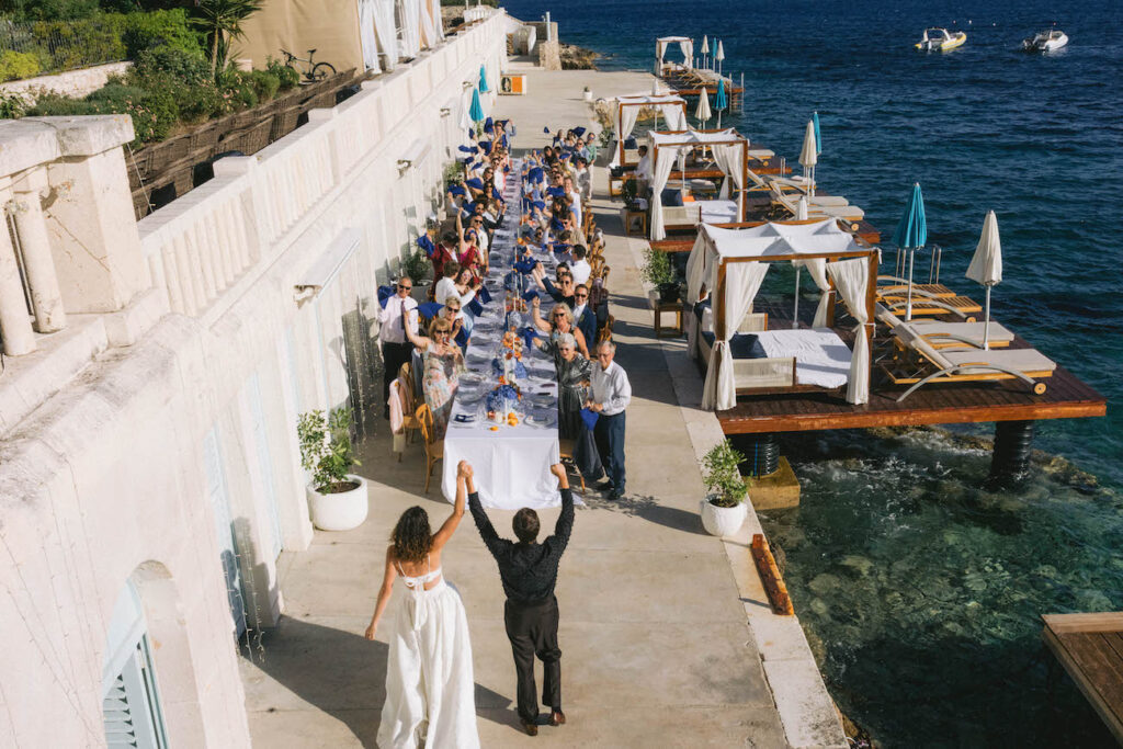A bride and groom walk toward a long outdoor dining table with guests seated on one side, next to cabanas and a waterfront.