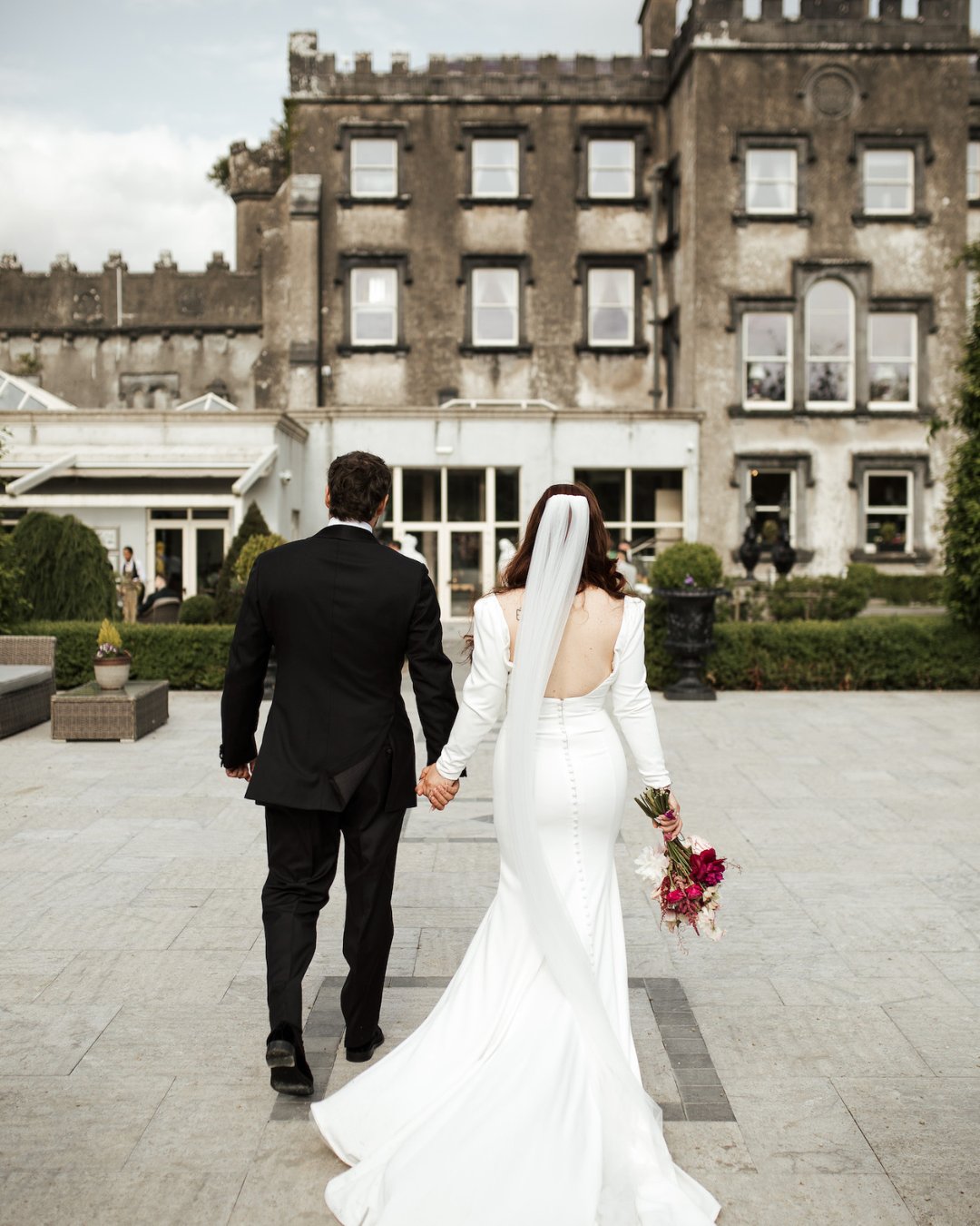A bride and groom holding hands walk towards a large stone building; the bride wears a white gown and veil, and the groom is in a black suit.