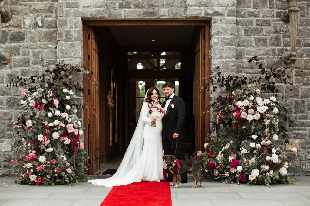 Bride and groom stand together with two dogs on a red carpet in front of a stone building, surrounded by large floral arrangements.