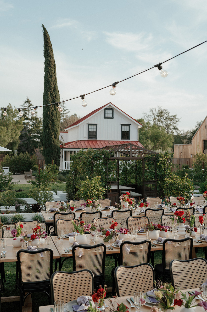 Outdoor event setup with long tables and chairs arranged for dining, decorated with colorful flowers, in front of a white house and garden, under string lights.
