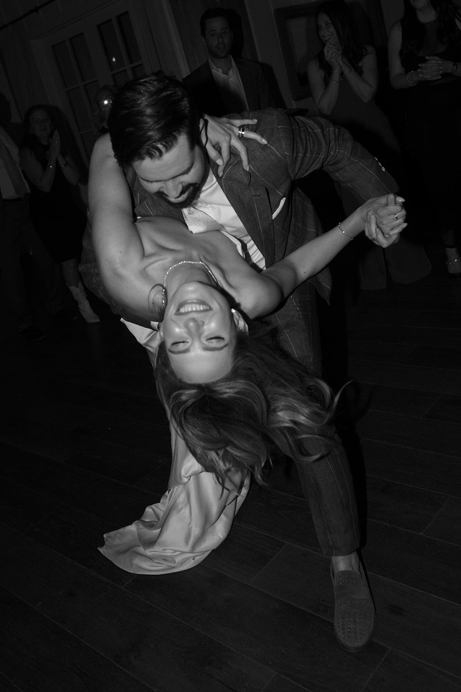 A man in a suit dips a smiling woman in a dress while dancing, surrounded by people clapping and watching in the background.