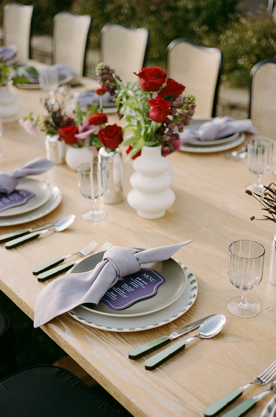 A wooden table set for a meal with plates, cutlery, glasses, folded napkins, and white vases holding red and purple flowers.