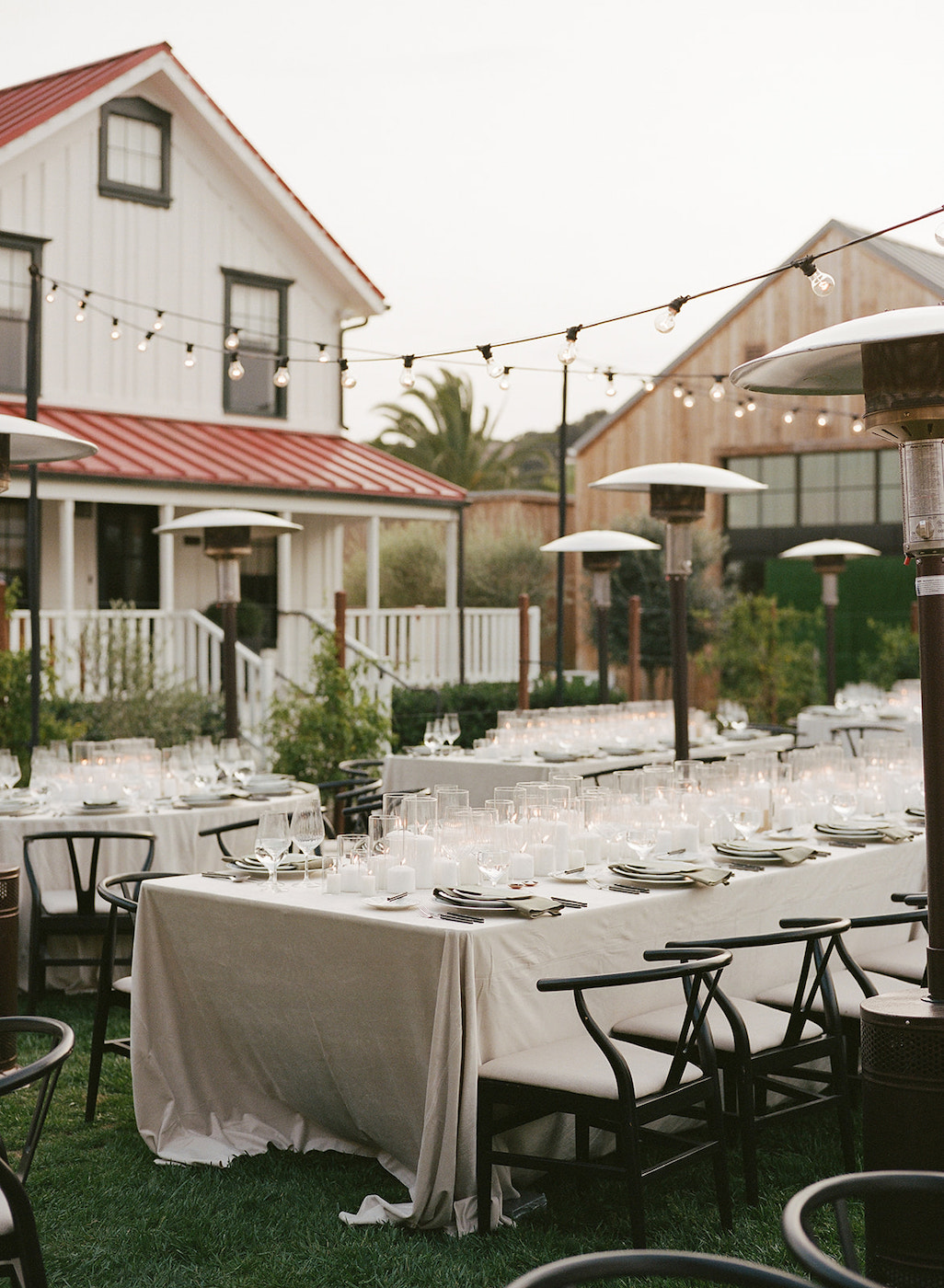 Outdoor dining setup with white tablecloths, black chairs, and string lights in a garden area between two farmhouse-style buildings.
