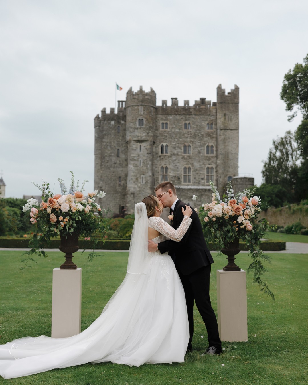 A bride and groom share a kiss outdoors in front of a stone castle, flanked by large floral arrangements on pedestals.