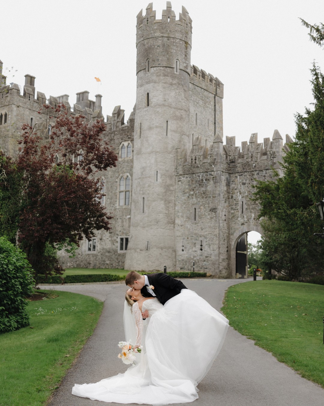 A bride and groom share a kiss as the groom dips the bride in front of a large stone castle on a lush, green pathway.