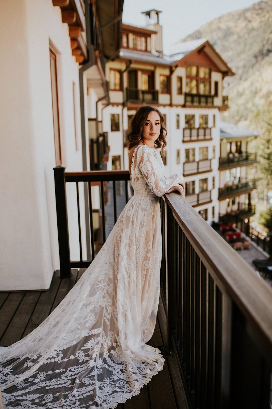 A woman in a long, lace wedding dress stands on a balcony overlooking a mountain resort with multi-story buildings in the background.
