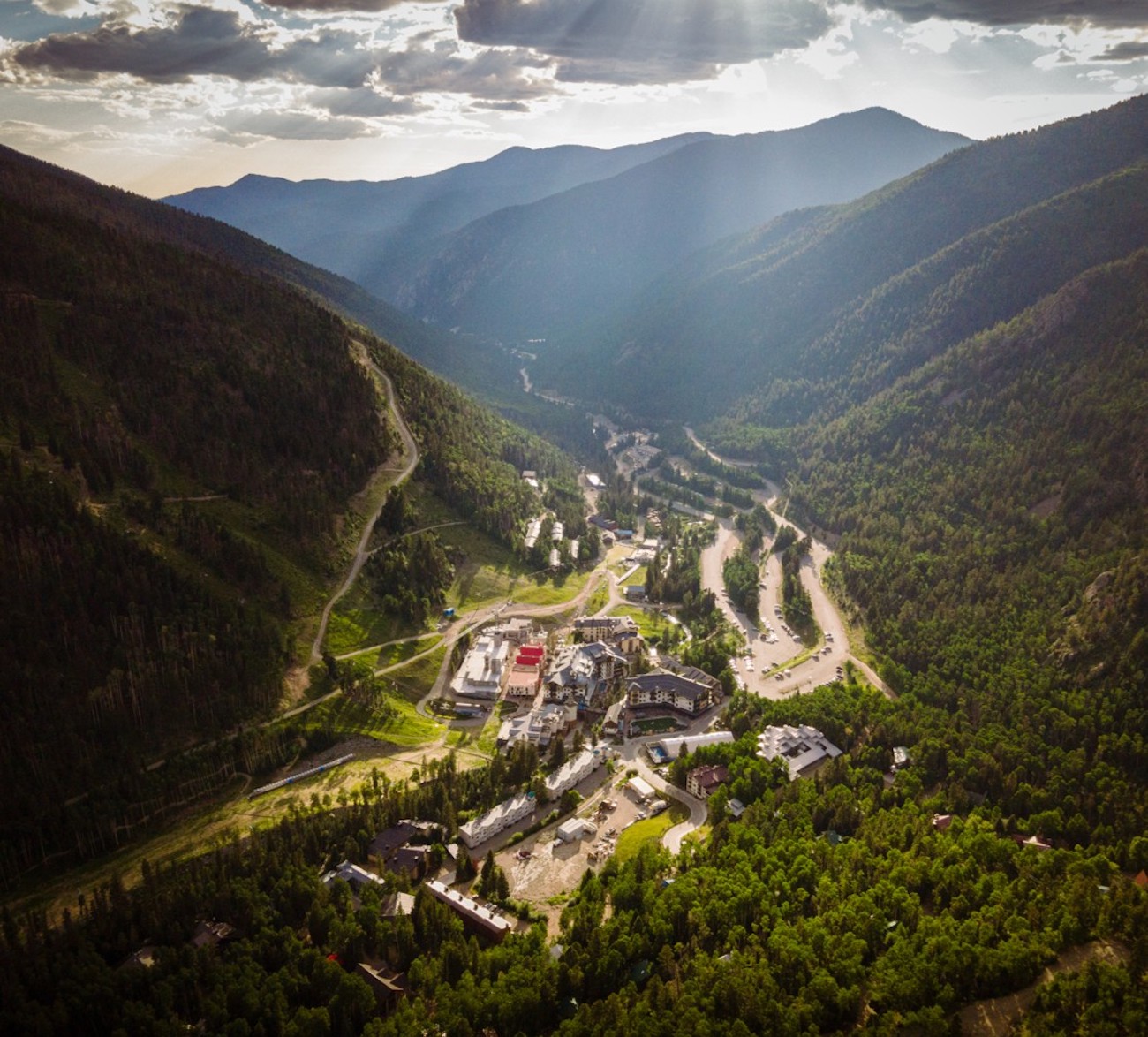 Aerial view of a small town nestled in a green mountain valley, with winding roads and scattered buildings, under a partly cloudy sky.