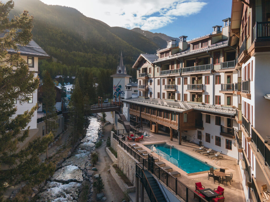 A hotel with balconies and an outdoor pool sits beside a stream and pine trees in a mountain valley, with a church and mountains in the background.
