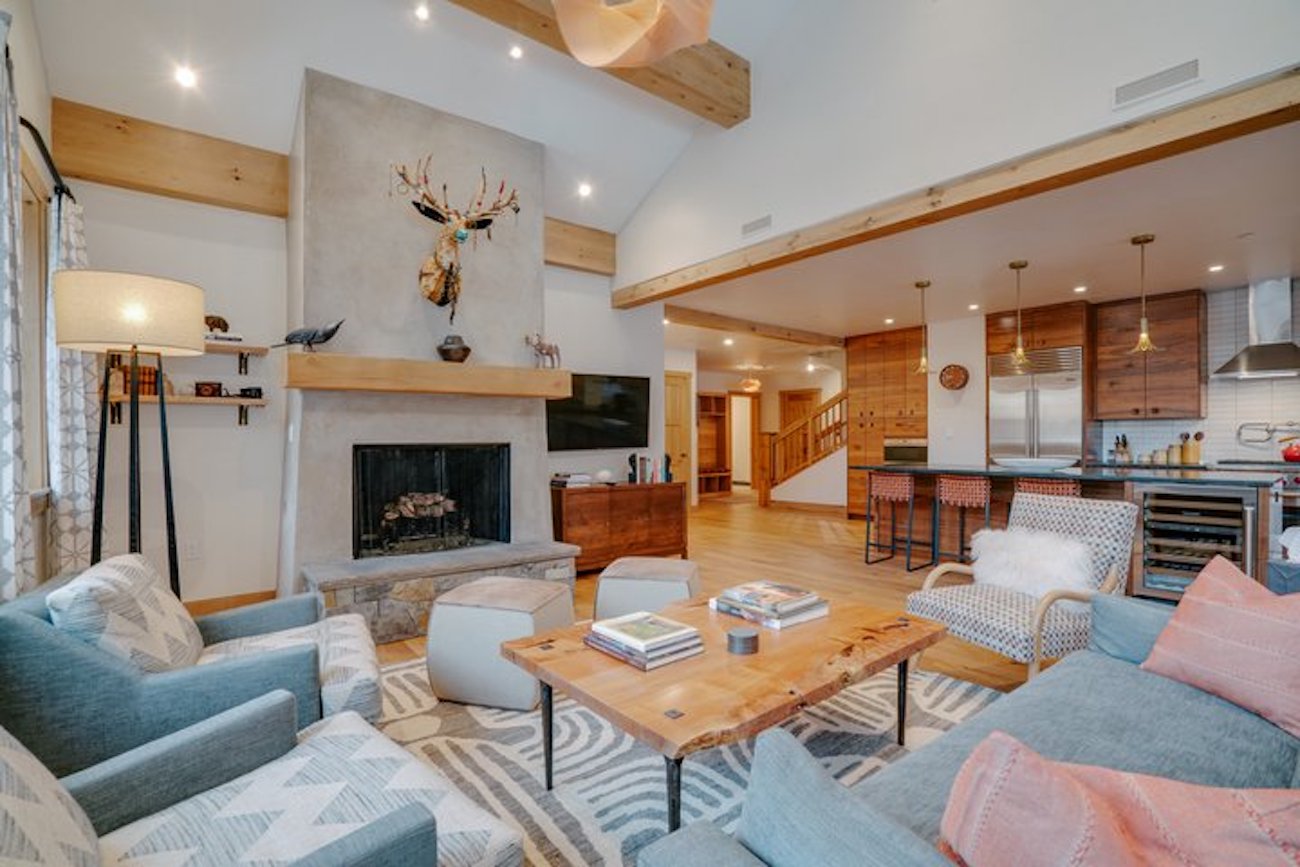 A spacious living room with a fireplace, mounted deer head, wooden beams, modern furniture, and an open kitchen in the background.