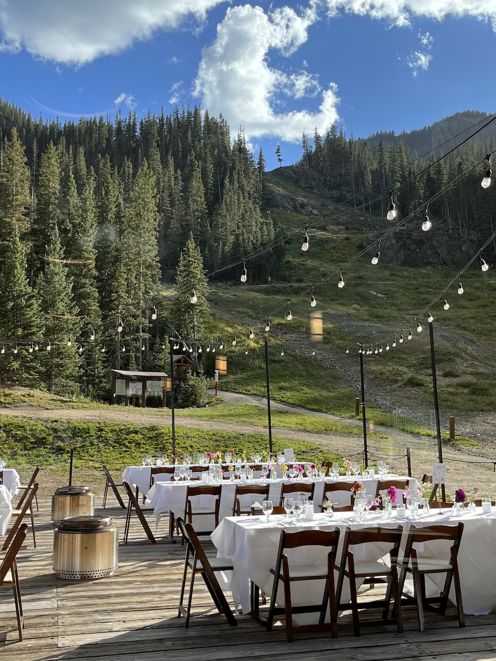 Outdoor dining setup with white tablecloths and wooden chairs on a wooden deck, surrounded by mountains, pine trees, and string lights under a partly cloudy sky.