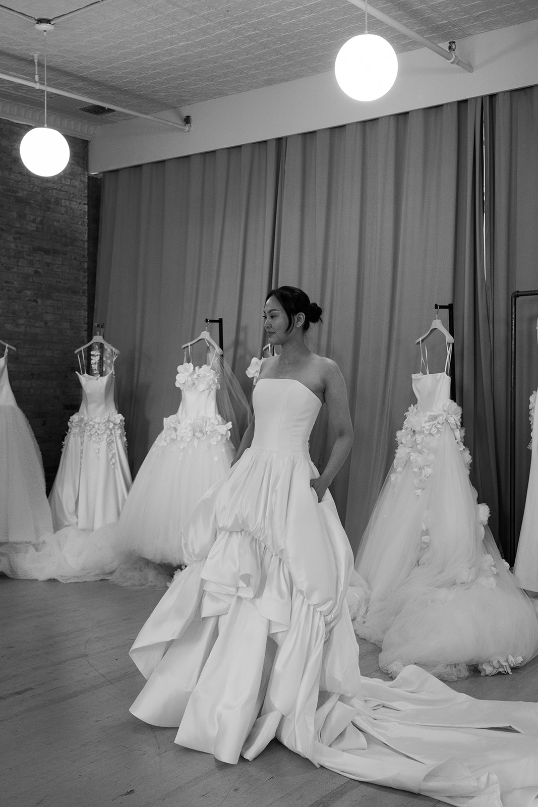A woman stands in a bridal shop wearing a strapless wedding dress, surrounded by mannequins displaying other wedding gowns.