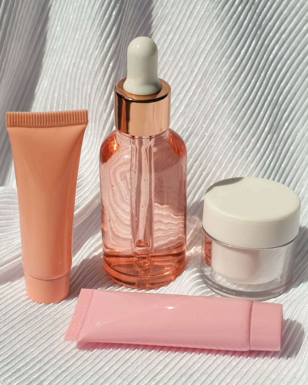 A set of skincare products including two tubes, a glass dropper bottle with a rose gold cap, and a small jar with a white lid, displayed on a white textured surface.