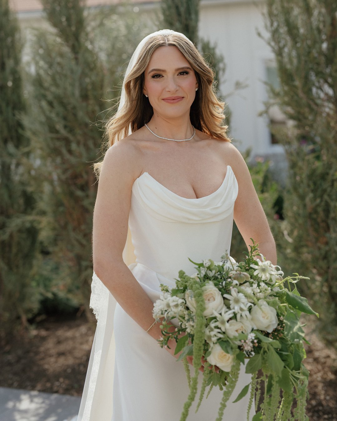 A woman in a white wedding dress holds a bouquet of white and green flowers, standing outdoors in front of tall greenery.