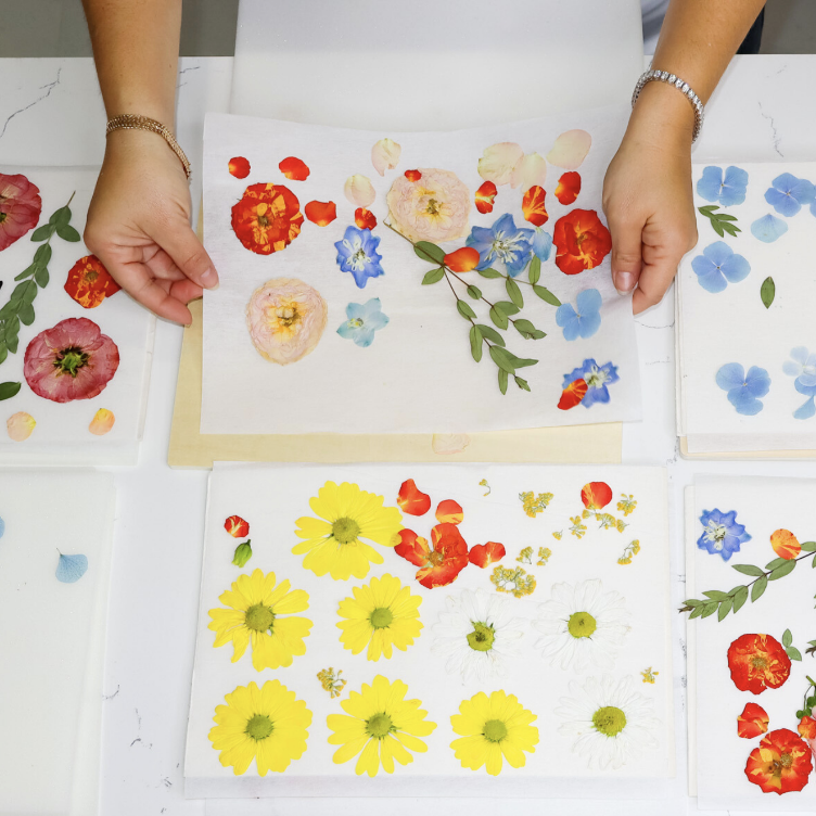A person arranges pressed flowers and leaves on white paper, with several sheets displaying different floral patterns on a table.