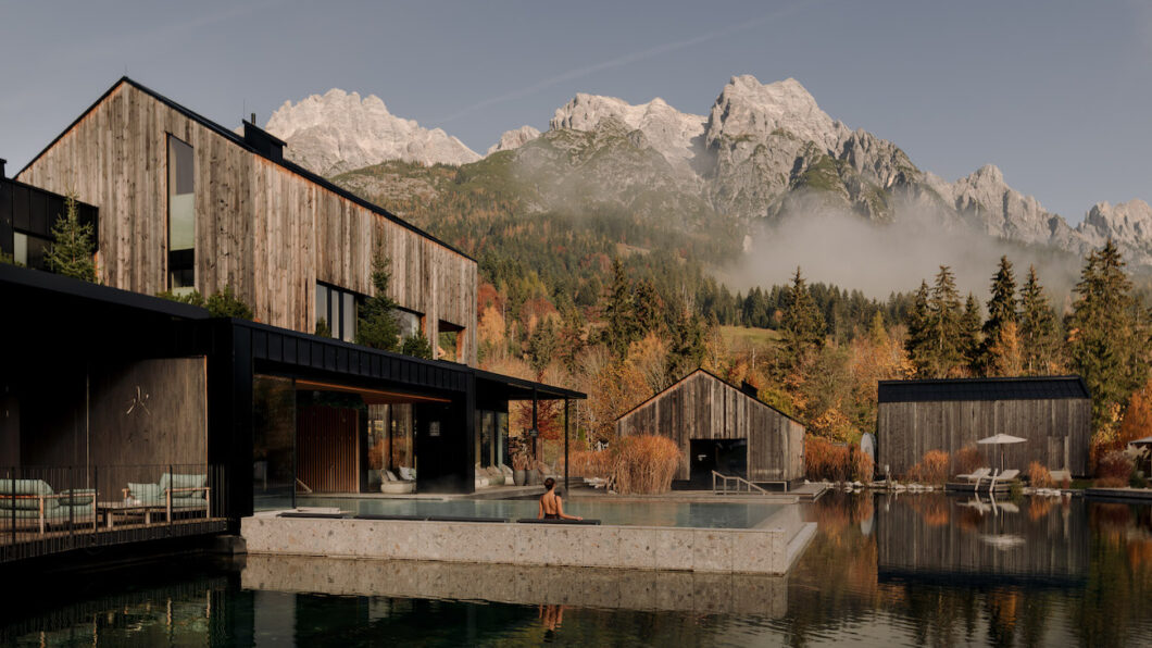 Modern wooden buildings surround a reflecting pool with a person swimming; mountains and trees are visible in the background under a clear sky.
