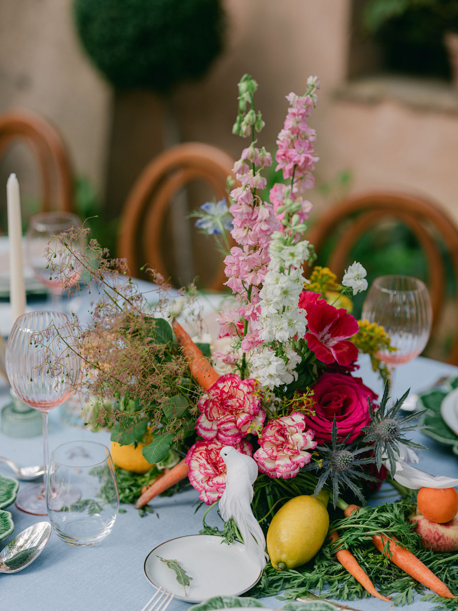 A floral centerpiece with pink, white, and green flowers, lemons, and carrots is arranged on a table set with glassware and plates.