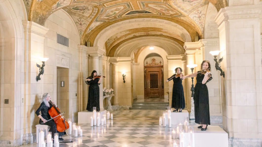 Four women in black dresses perform with string instruments in an ornate hall with arched ceilings, columns, and candles on the checkered floor.
