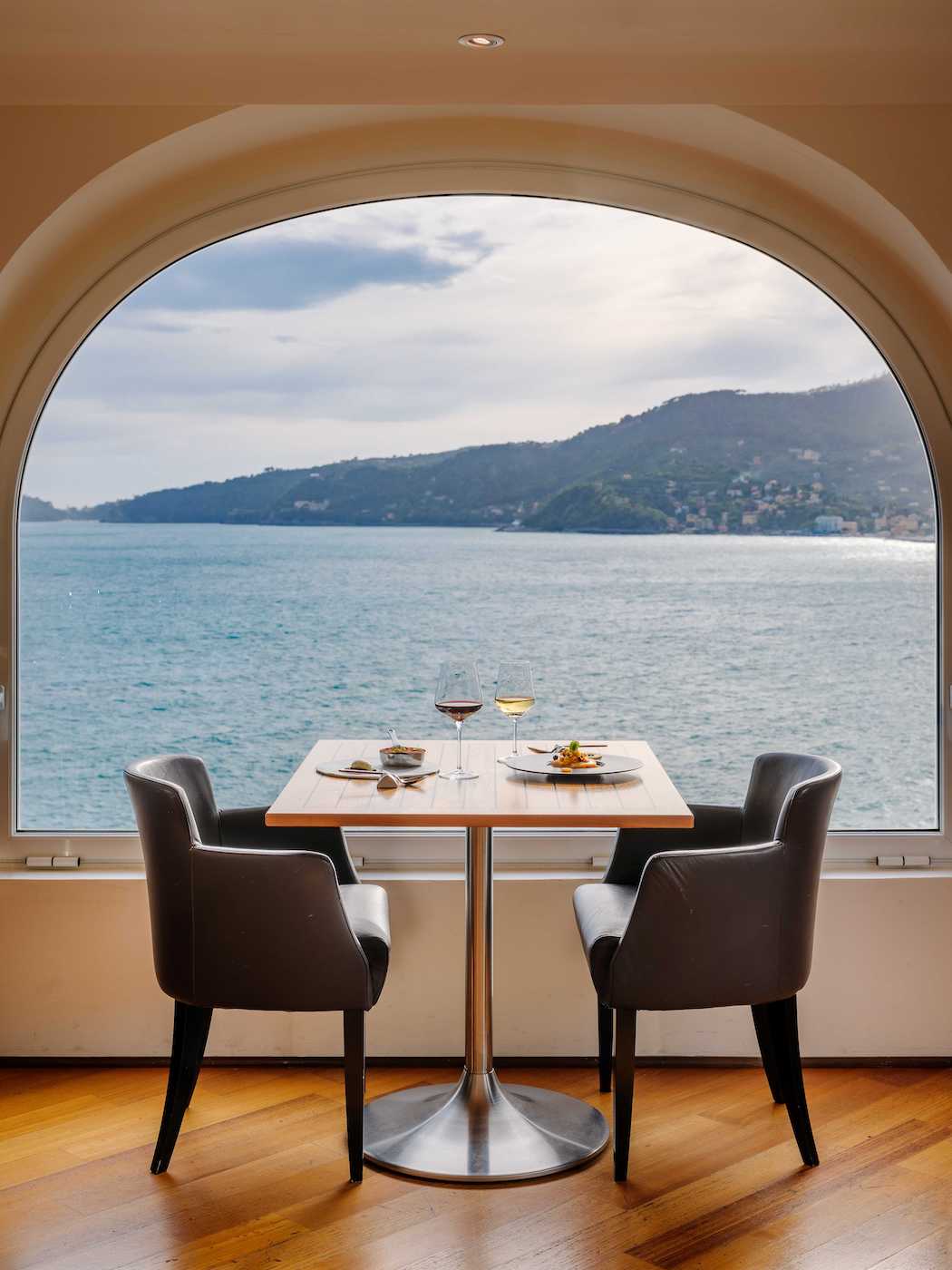 A dining table set for two with plates and glasses sits by a large arched window overlooking a body of water and distant hills.