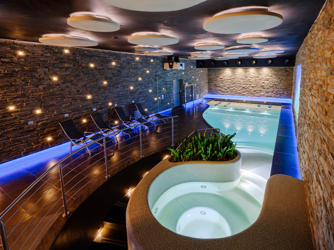 Indoor spa area with a modern pool, hot tub, lounge chairs, stone walls, soft ceiling lights, and accent lighting along the floor and pool edges.