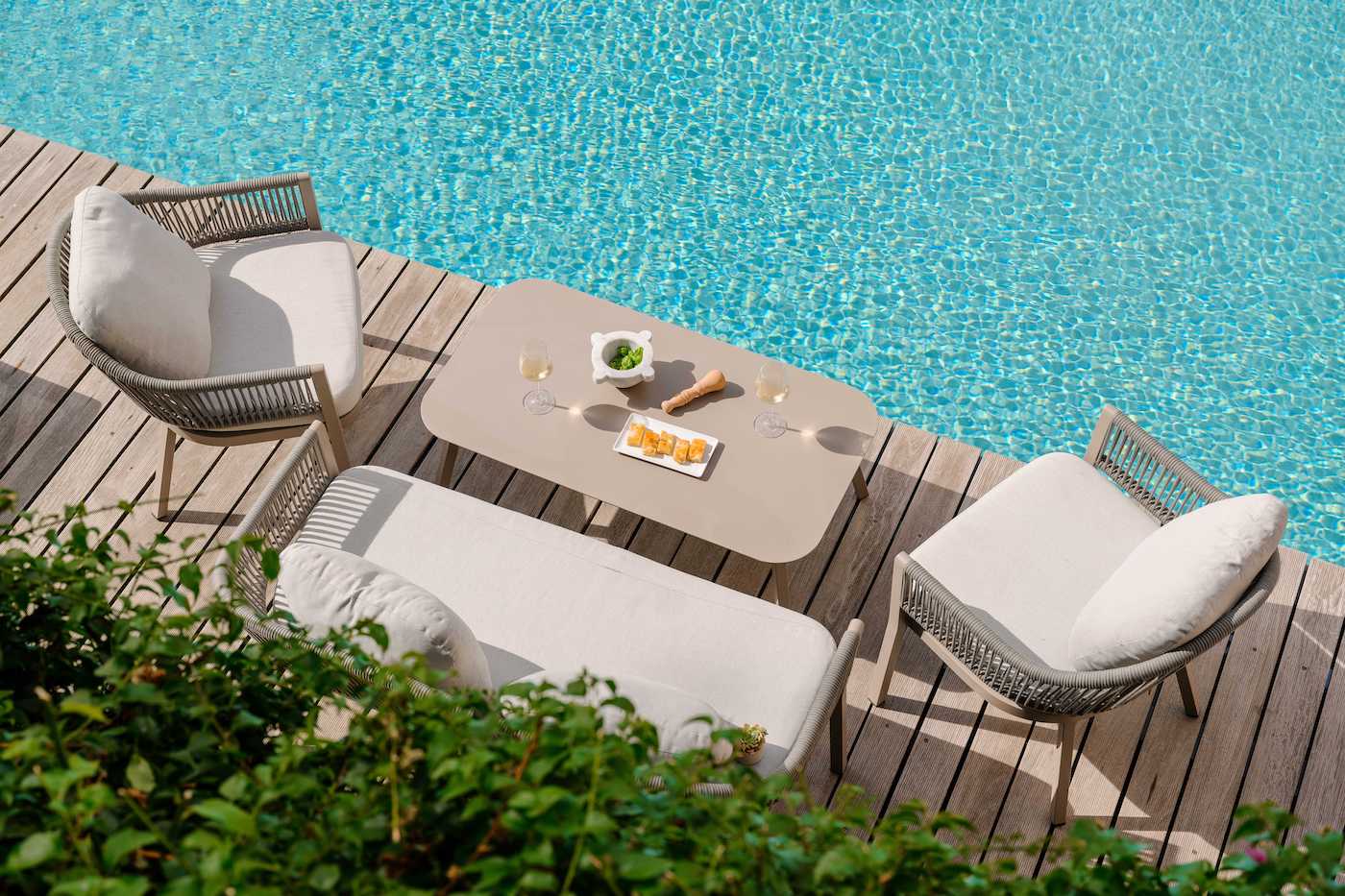 Outdoor poolside seating area with two chairs, a bench, and a table set with snacks and drinks on a wooden deck beside a clear blue swimming pool.