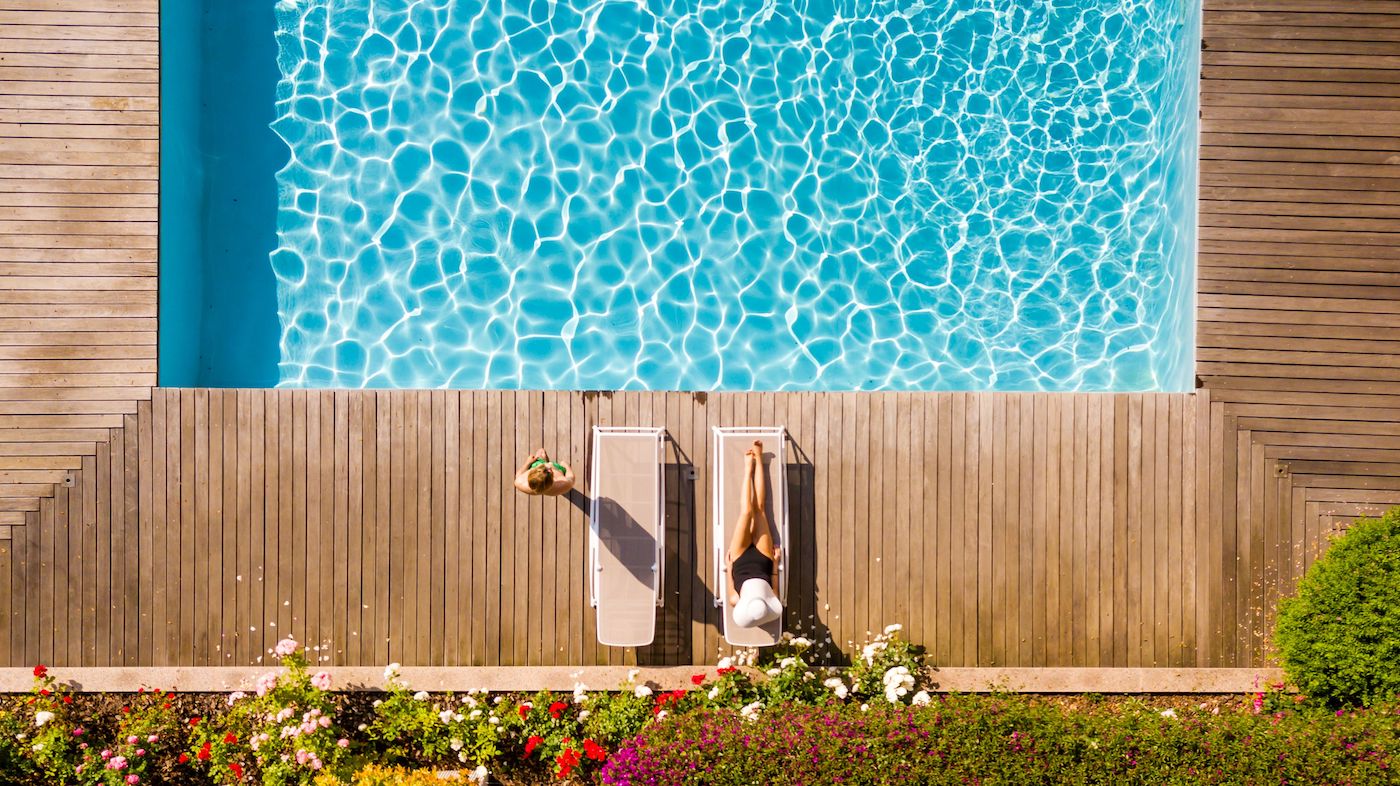 A person is lying on a lounge chair next to a pool, with their legs raised on another chair. The pool water is clear and surrounded by wooden decking and flowers.