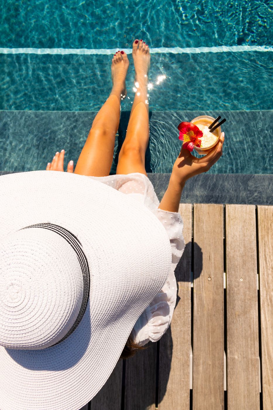 Person wearing a large white sunhat sits by a pool with legs in the water, holding a tropical drink, on a wooden deck.