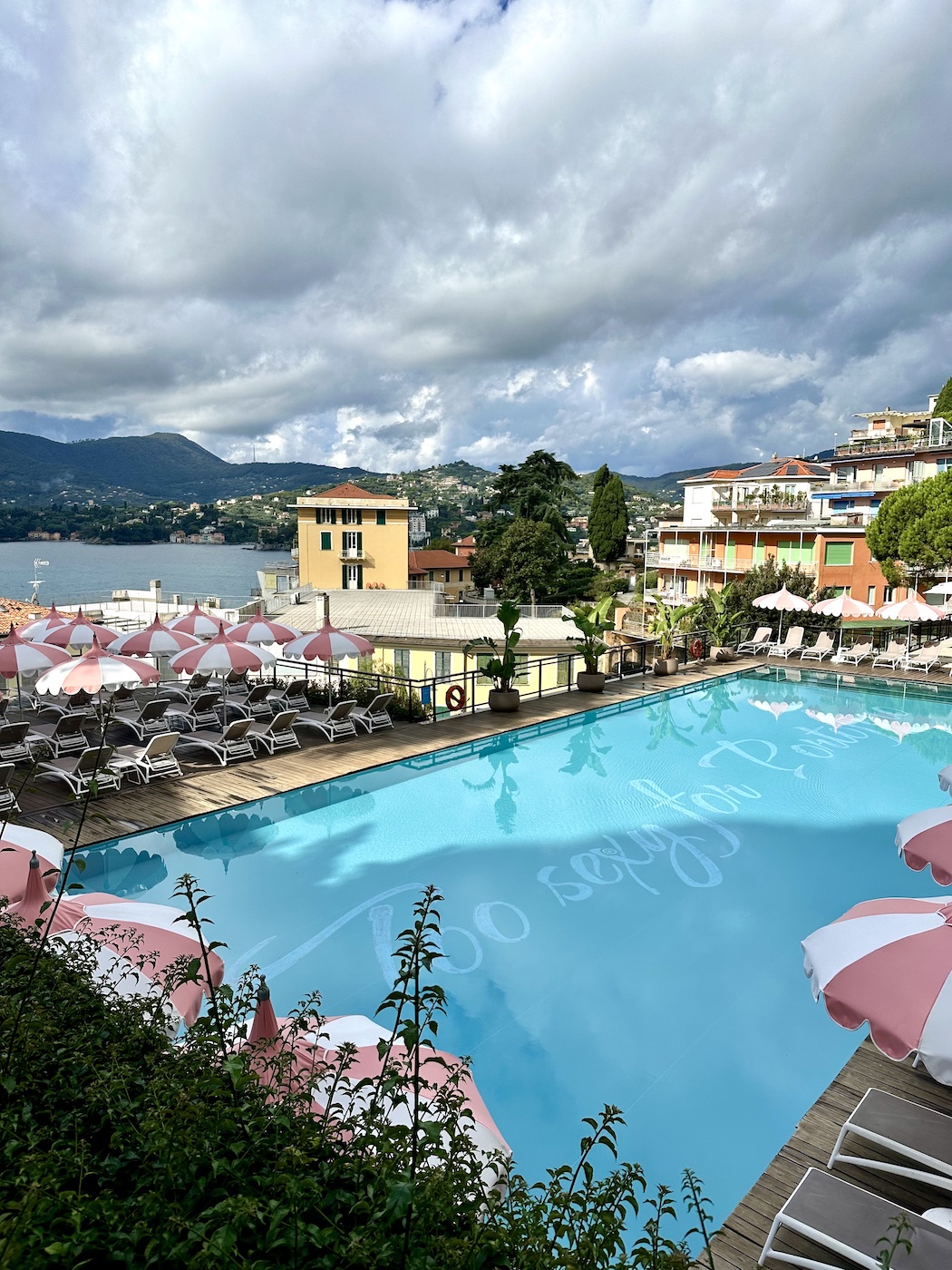 Large outdoor swimming pool surrounded by pink and white lounge chairs and umbrellas, with buildings, mountains, and a lake in the background under a cloudy sky.