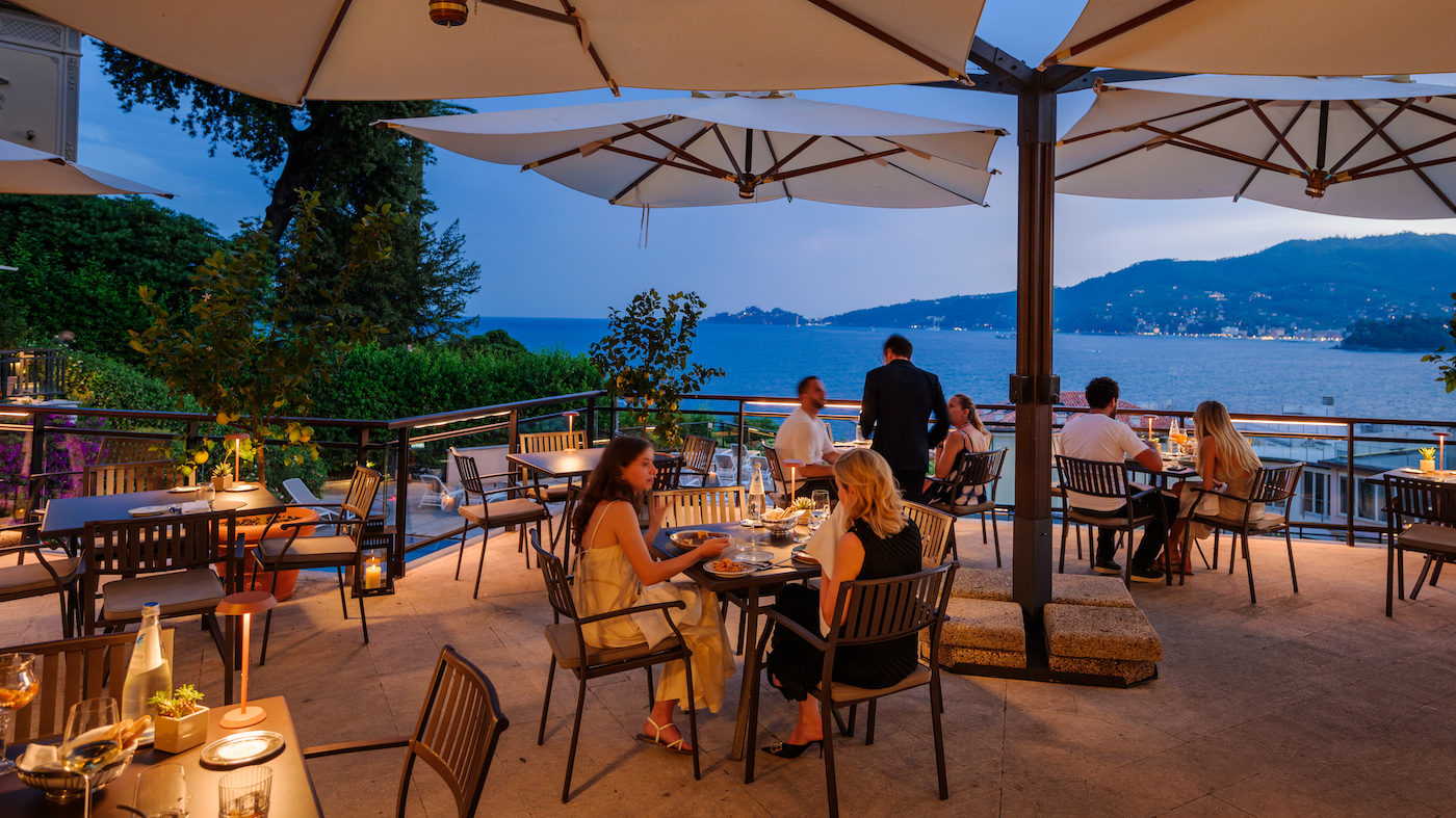 People dine at outdoor restaurant tables under large umbrellas in the evening, overlooking a body of water and distant hills.