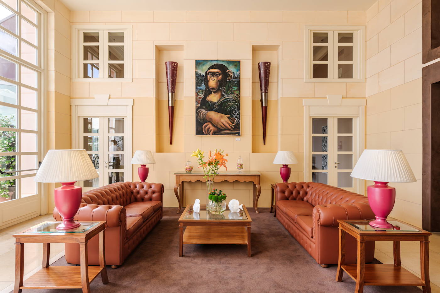 A formal living room with two brown leather sofas, a wooden coffee table, pink lamps, and a framed portrait on the wall above a console table with flowers.