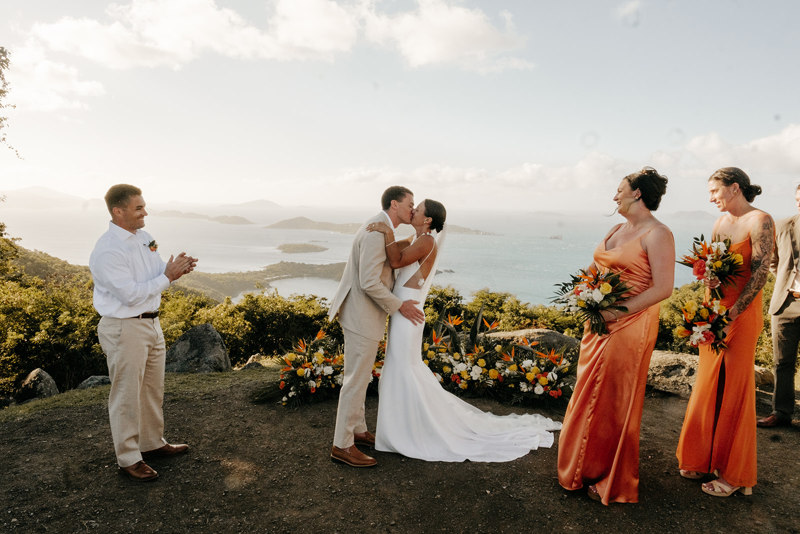 A bride and groom kiss outdoors at their wedding ceremony, with two attendants standing nearby and scenic ocean views in the background.