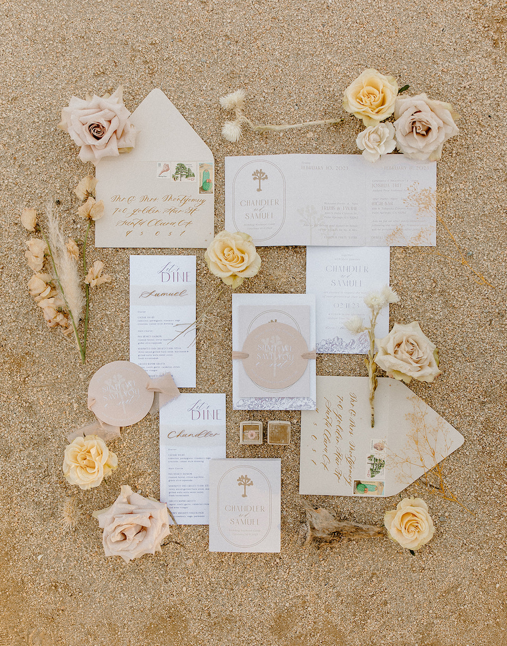 Flat lay of wedding stationery, envelopes, menus, and invites arranged on sand, accented with neutral-toned roses and dried floral elements.