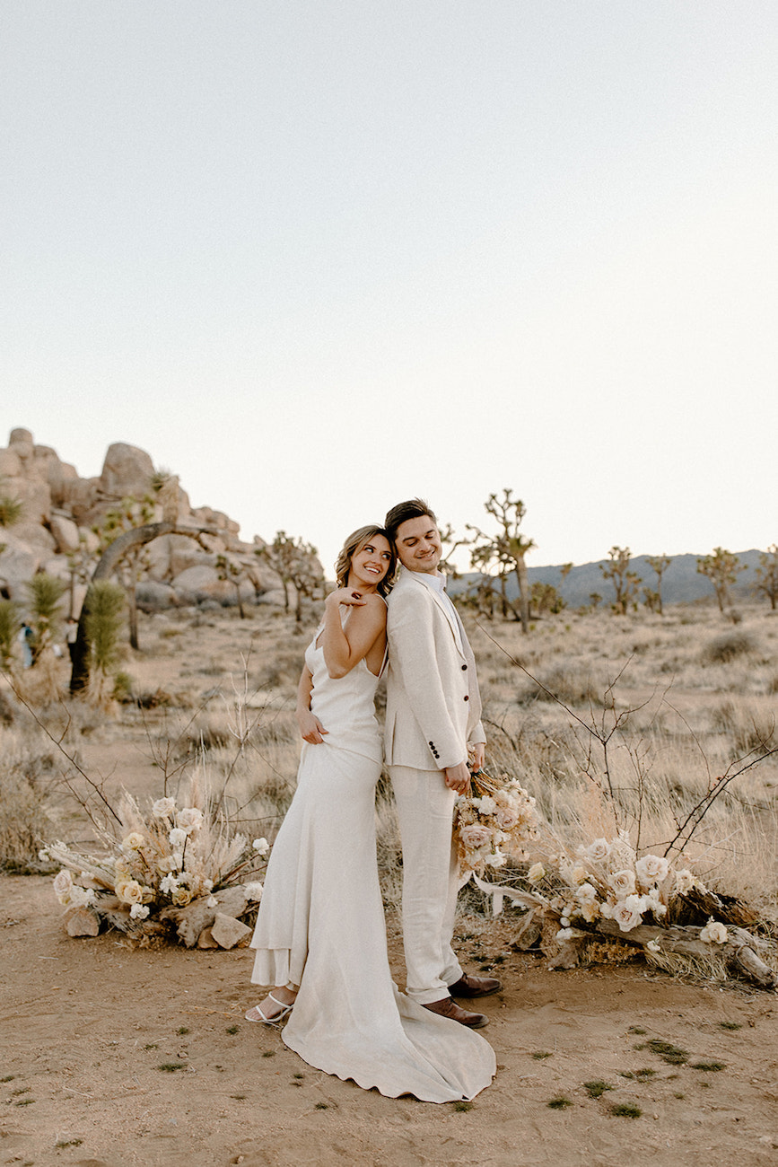 A bride and groom in white attire stand back to back, smiling, in a desert landscape with dried floral arrangements and rocky hills in the background.