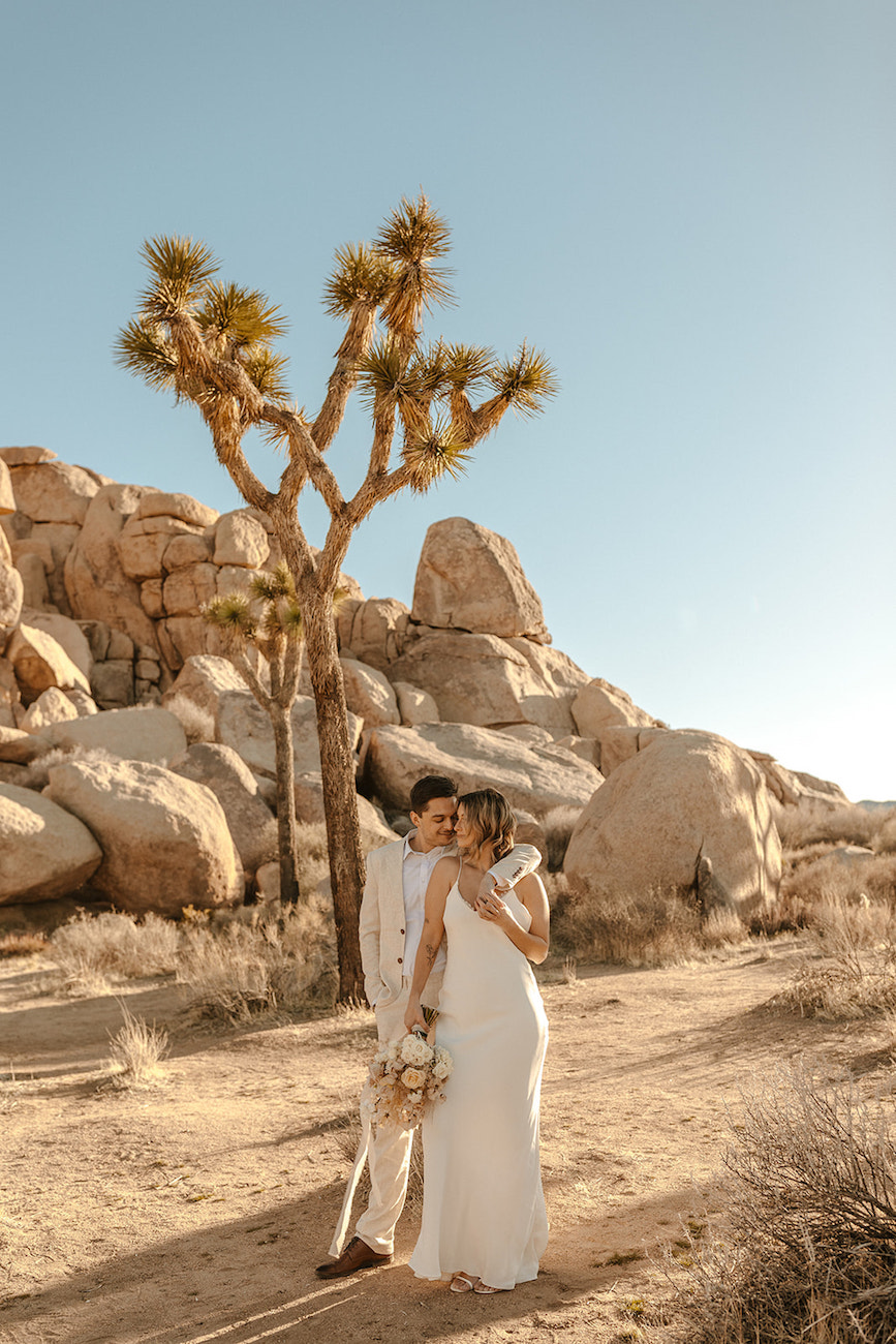 A couple in wedding attire stands close together in a desert landscape with large rocks and a Joshua tree in the background under a clear sky.