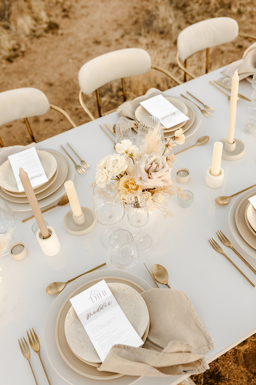 A table set outdoors with neutral-toned plates, gold cutlery, beige napkins, menus, candles, and a dried floral centerpiece.
