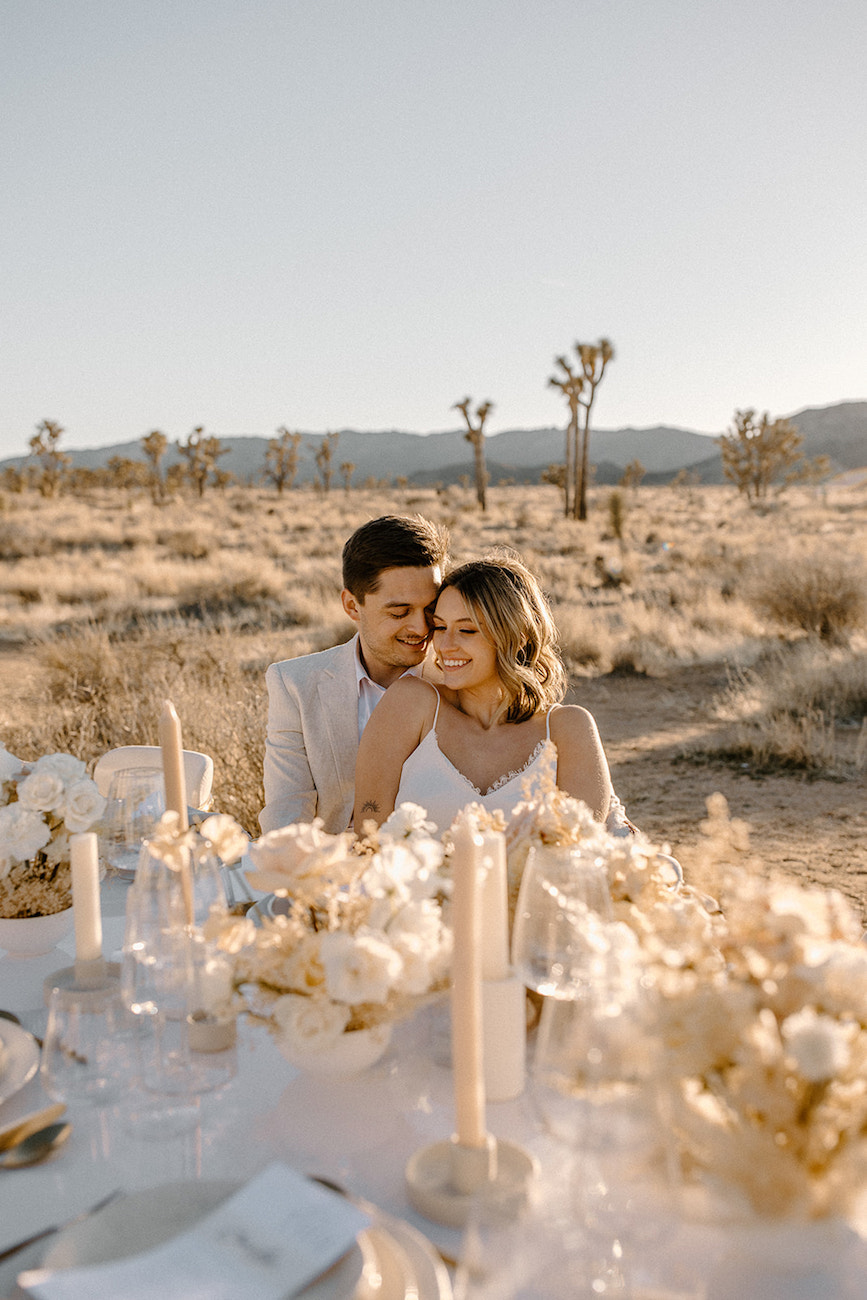 A couple sits closely together at an elegantly decorated outdoor table in a desert landscape, surrounded by flowers and candles, with mountains in the background.