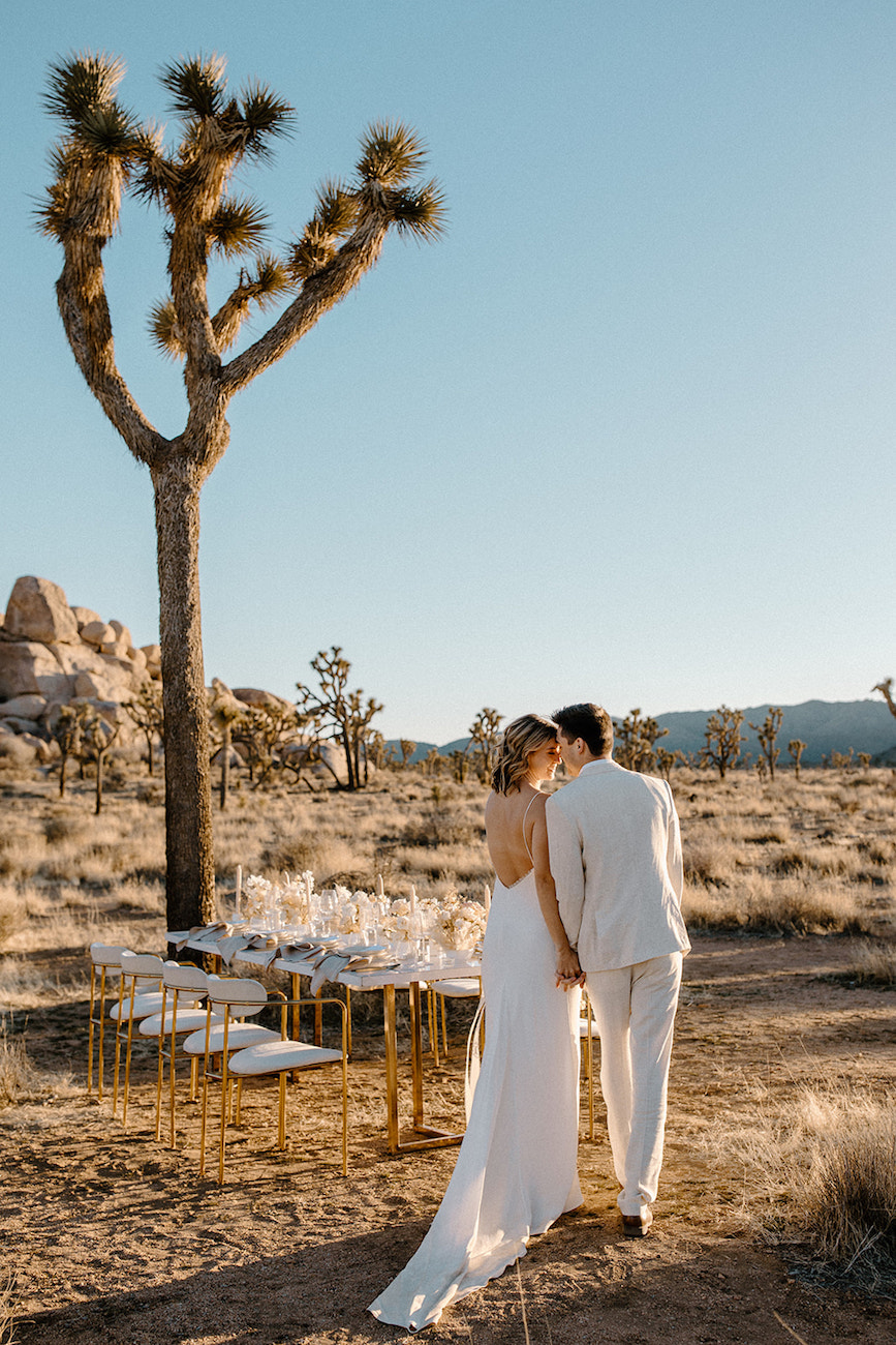 A couple in white wedding attire stands close together in a desert landscape with Joshua trees beside a set outdoor dining table.