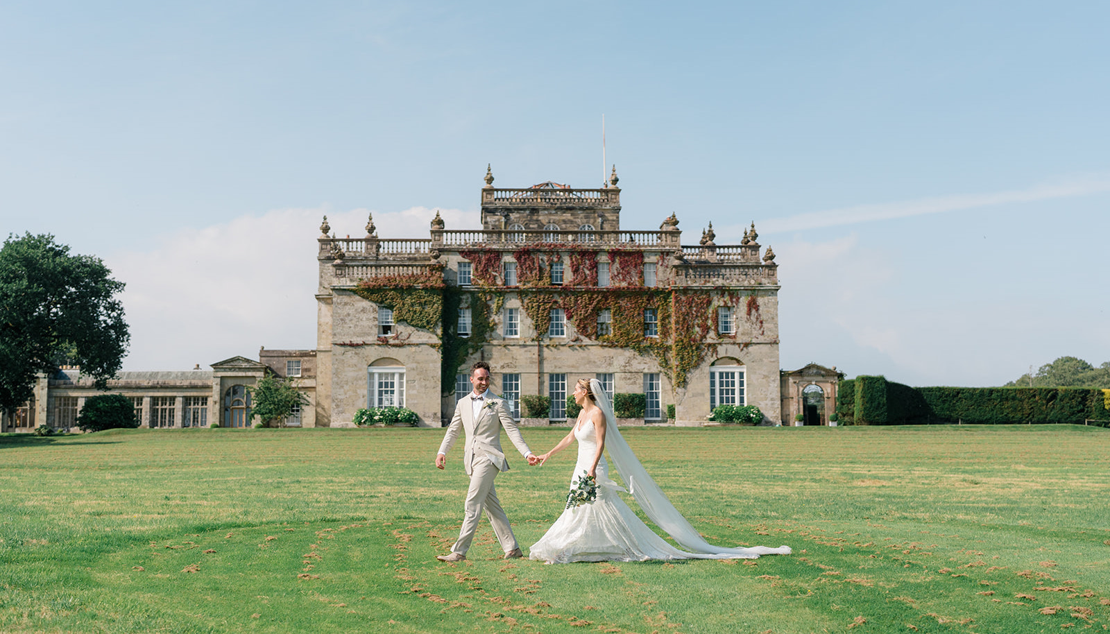 A bride and groom walk hand in hand on a lawn in front of a large, historic stone mansion covered in ivy.