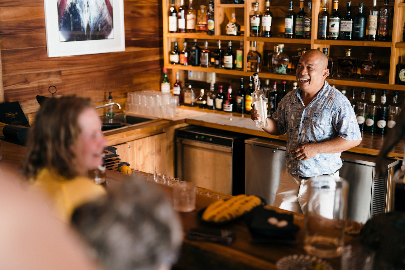 A man stands behind a wooden bar holding a drink, with shelves of liquor bottles behind him and people seated at the bar.