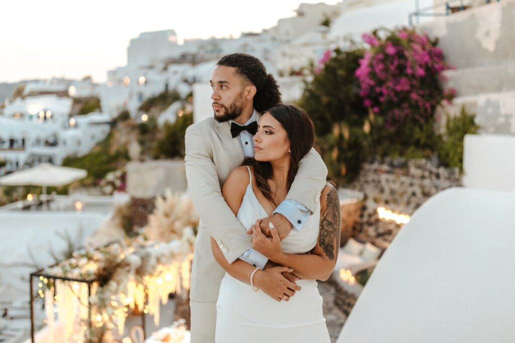 A man in a light suit embraces a woman in a white dress outdoors, with white buildings and pink flowers in the background.