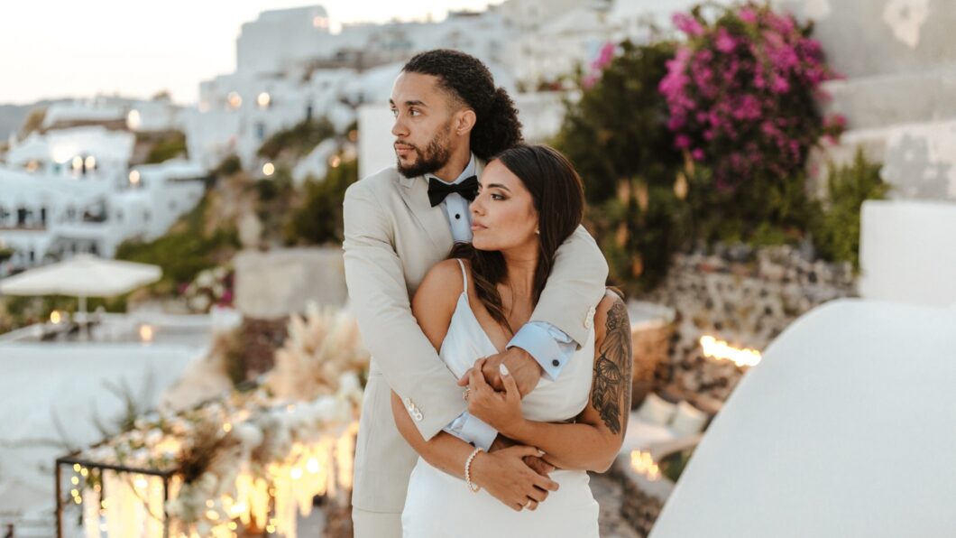 A man in a light suit embraces a woman in a white dress outdoors, with white buildings and pink flowers in the background.