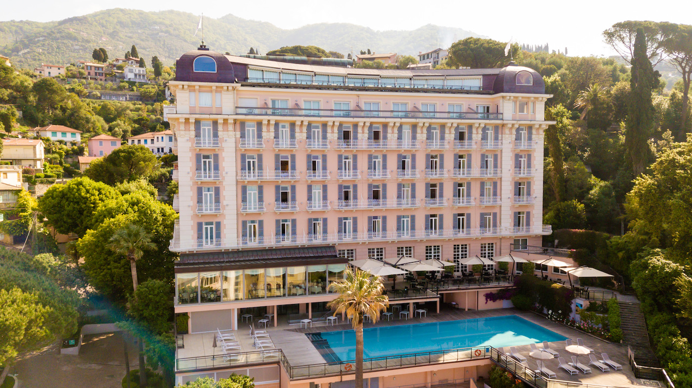 A large, light-colored hotel building with multiple floors and balconies overlooks an outdoor swimming pool, surrounded by trees and hills in the background.