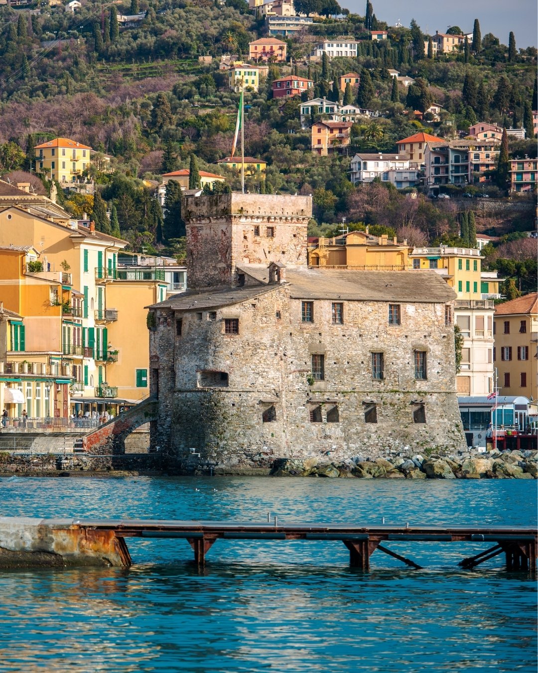 A stone castle-like building sits by the waterfront, surrounded by colorful houses and hills with trees and more buildings in the background.