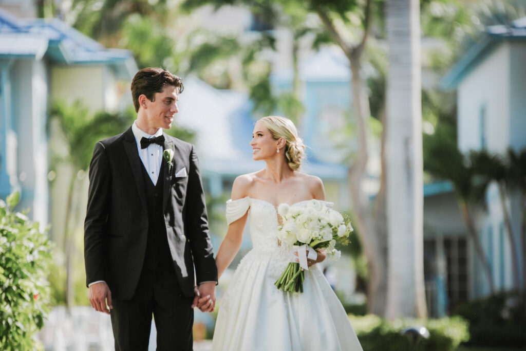 A bride and groom in formal wedding attire hold hands and look at each other while walking outdoors, surrounded by greenery and buildings.